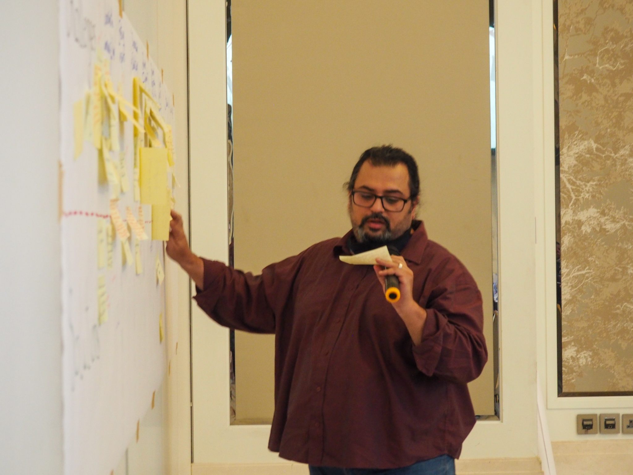 Man speaking on microphone, reading a note and his other hand pointing at a board with sticky notes